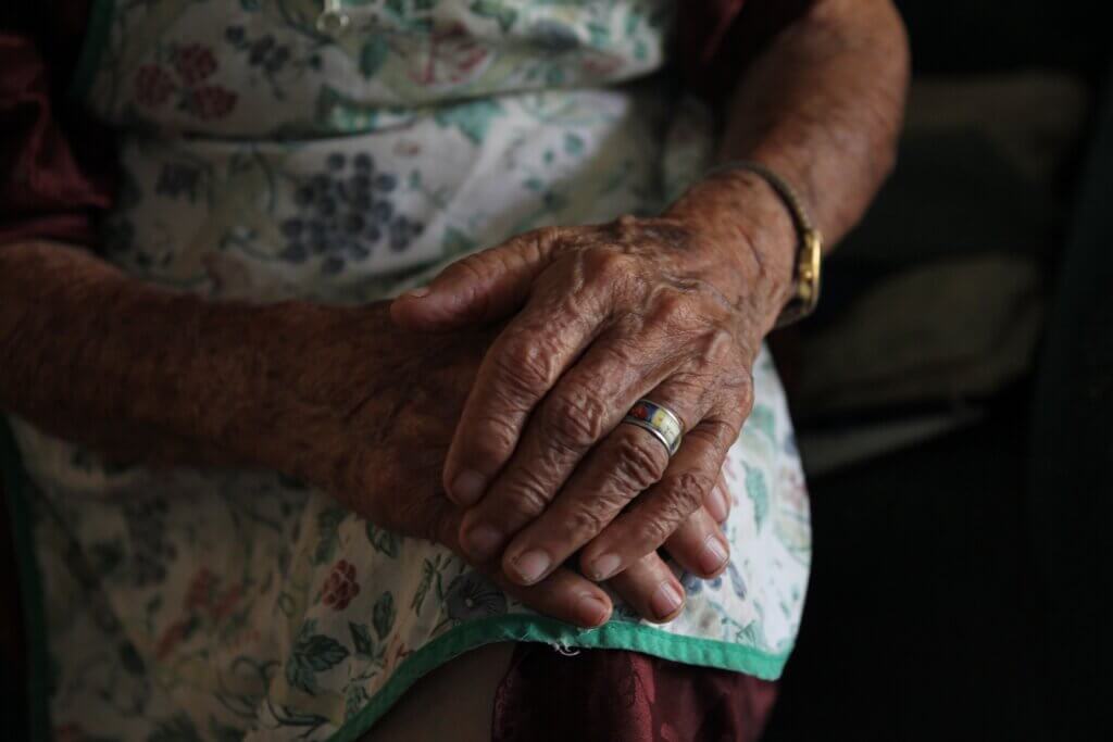 An elderly wrinkled woman's hands are clasped in the soft focus of sunlight.