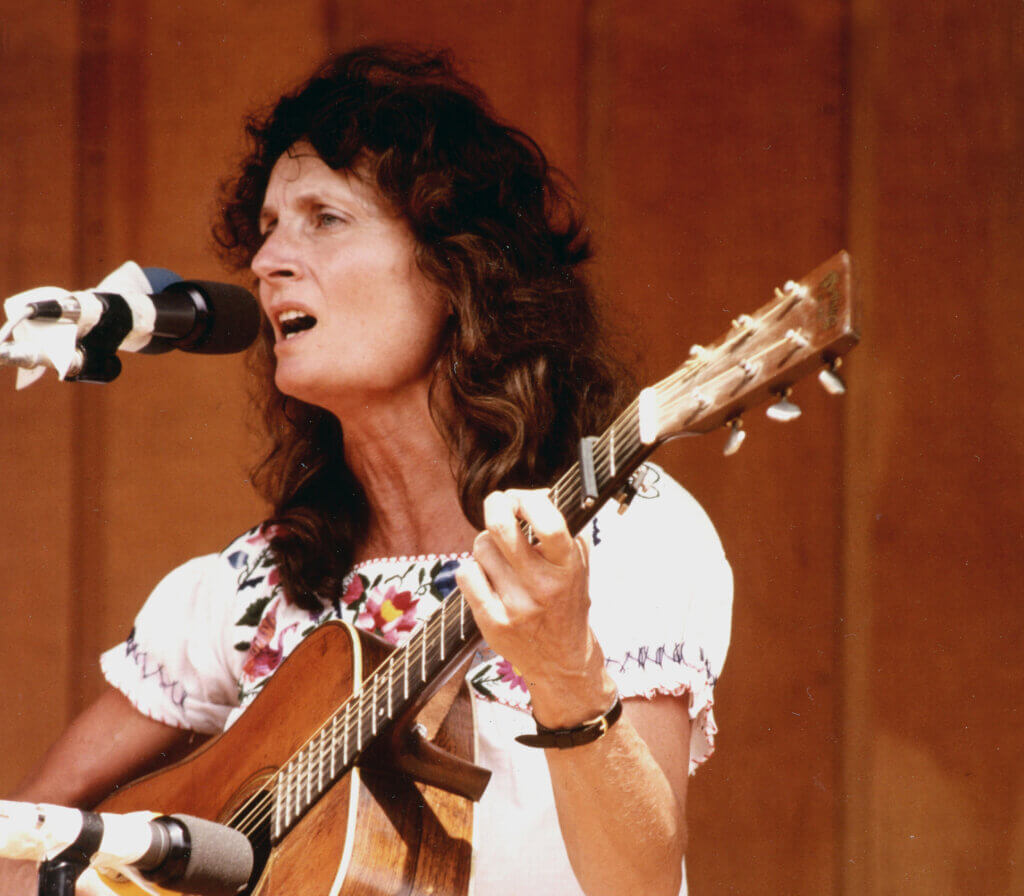 Alice Gerrard sings while holding and playing guitar.