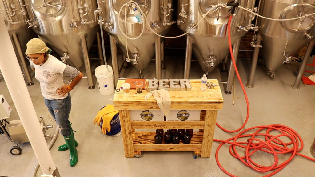 Overhead shot of inside of brewery. Black woman stands to side with hands on hips looking deep in thought. Metal sign "BEER" on wooden table in middle of the room to her left.
