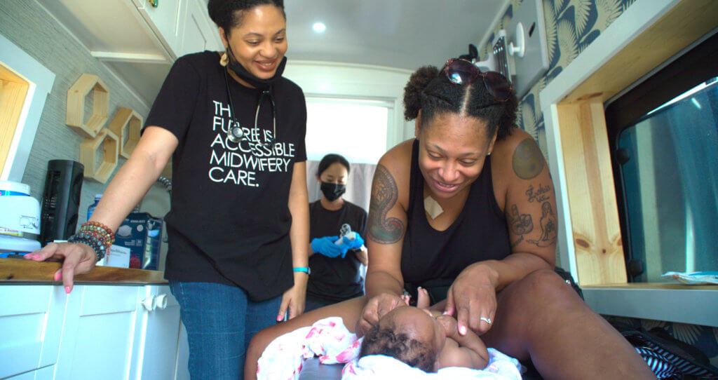 Tanya Smith-Johnson smiles as she stands over the shoulder of her client during a postpartum visit in a mobile midwifery van in Hawaii. Her client, a Black mother with tatoos, sits on an exam table and smiles at her baby, a newborn with curly tufts of black hair. Tanya has a stethoscope around her neck and wears a black T-shirt emblazoned with the slogan "THE FUTURE IS ACCESSIBLE MIDWIFERY CARE."