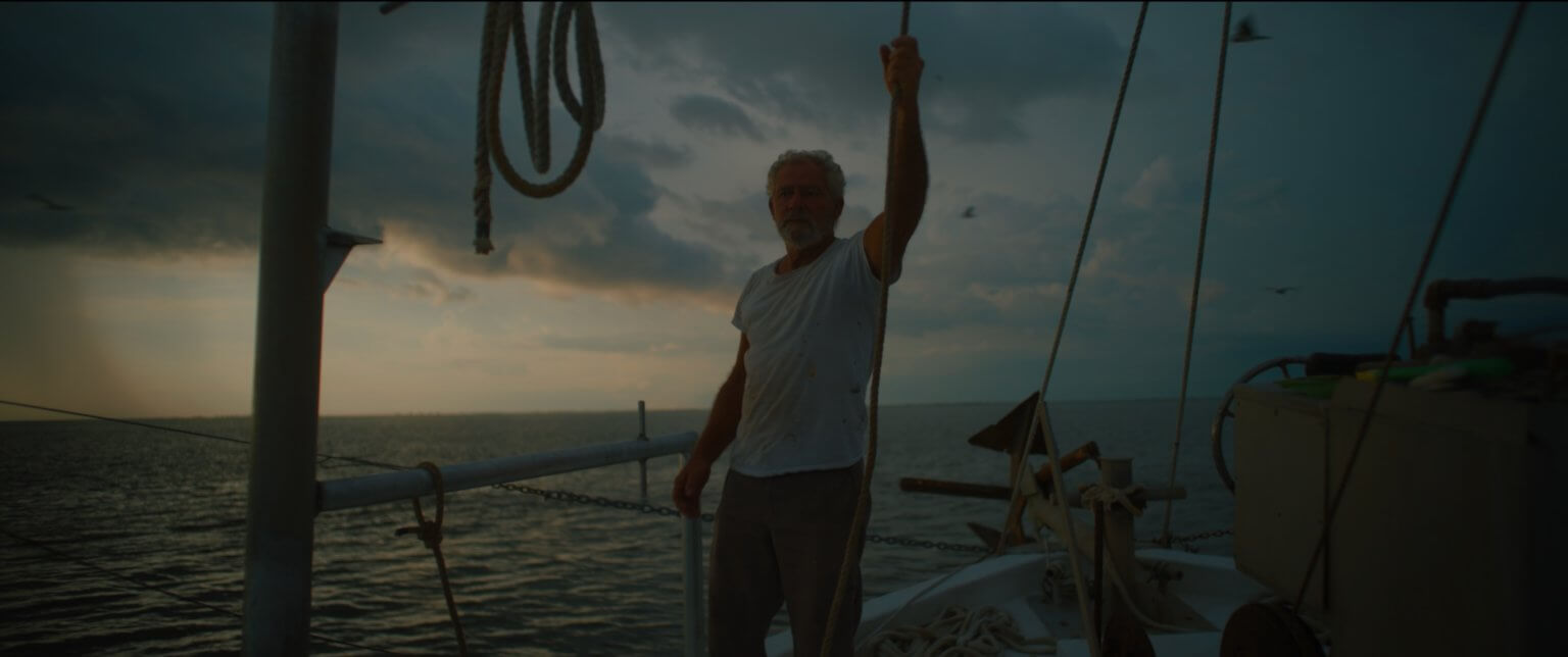 An older shrimper with grey hair stands on the bow of his boat in the Mississippi River. Behind him is a big, stormy sky.