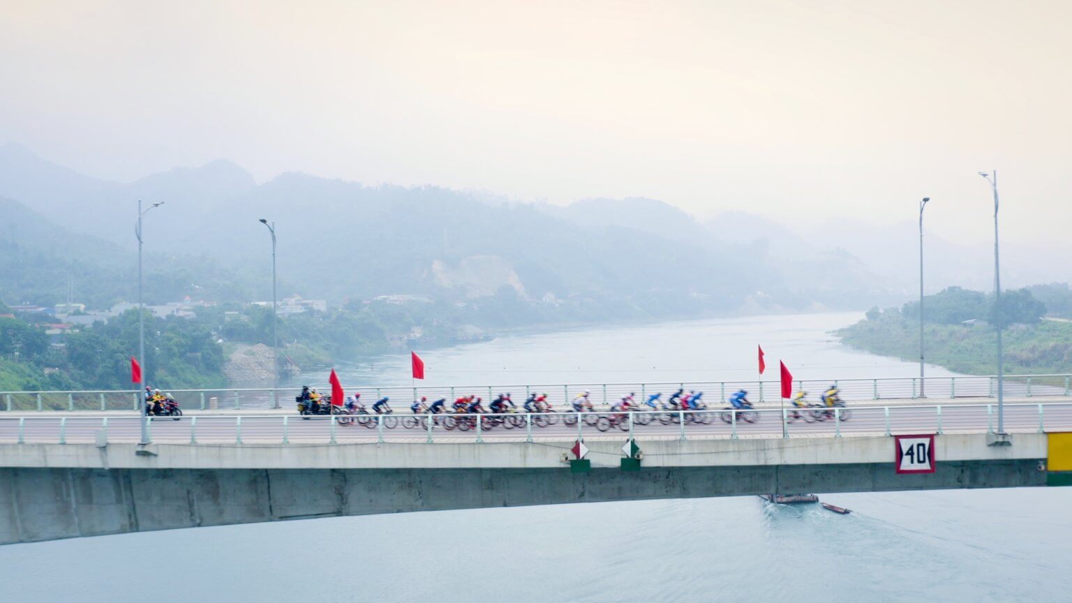 Aerial image showing a bridge running across the frame. The bridge is above a wide river and soft blue mountains are in the distance. On the bridge is a peloton of cyclists riding from left to right across the bridge.
