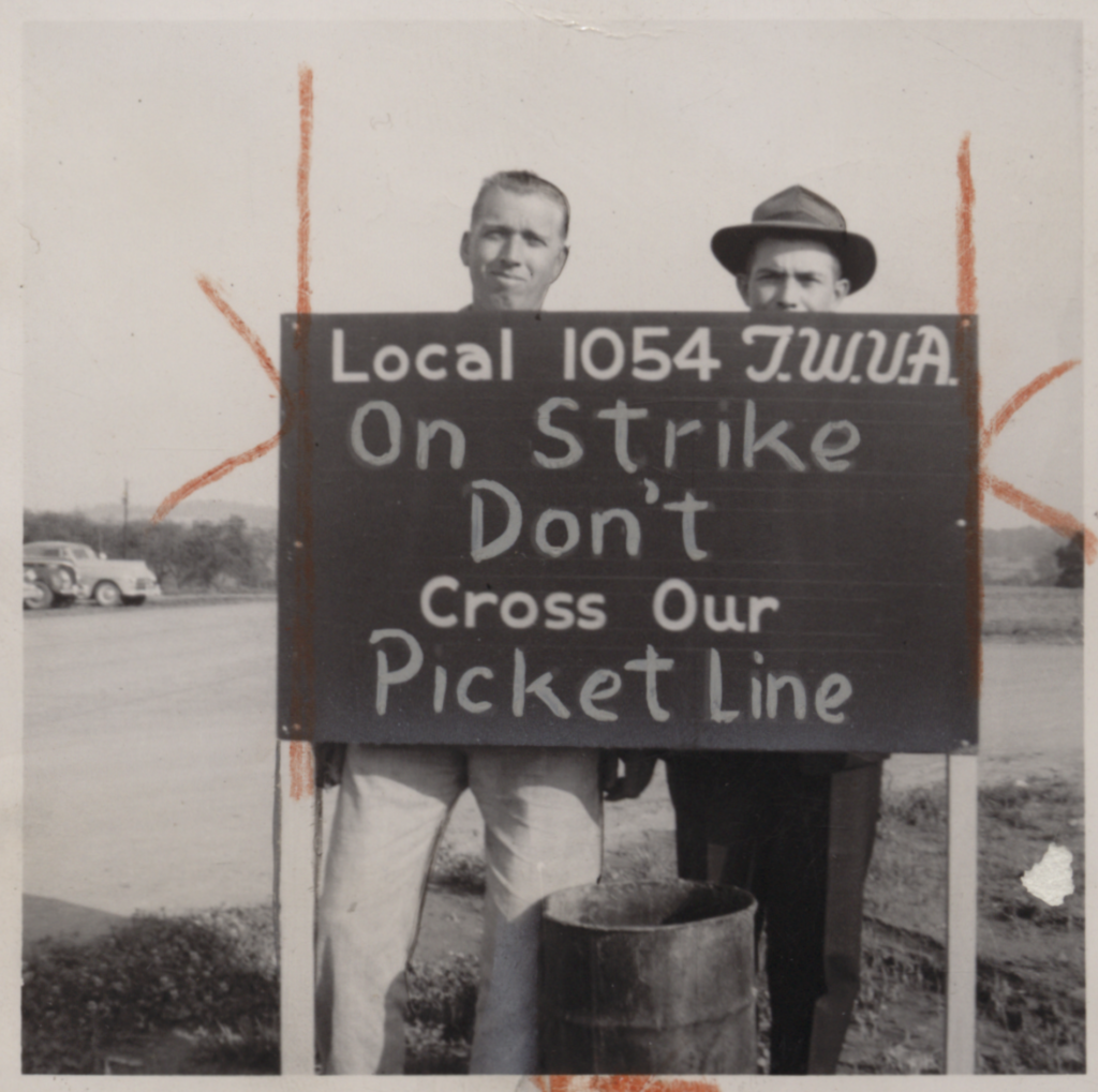 Two men stand behind a sign that reads "Local 1054 TWUA on strike. Don't cross our picket line."