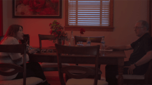 A young woman and her uncle sit at opposite ends on a dining table in a room basked in red light. They are in the middle of a conversation, she with her phone in hand, him with his hand on his gun pocket.