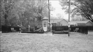 Black and white photo of an empty playground, on a rainy day. Surrounded by trees and grass