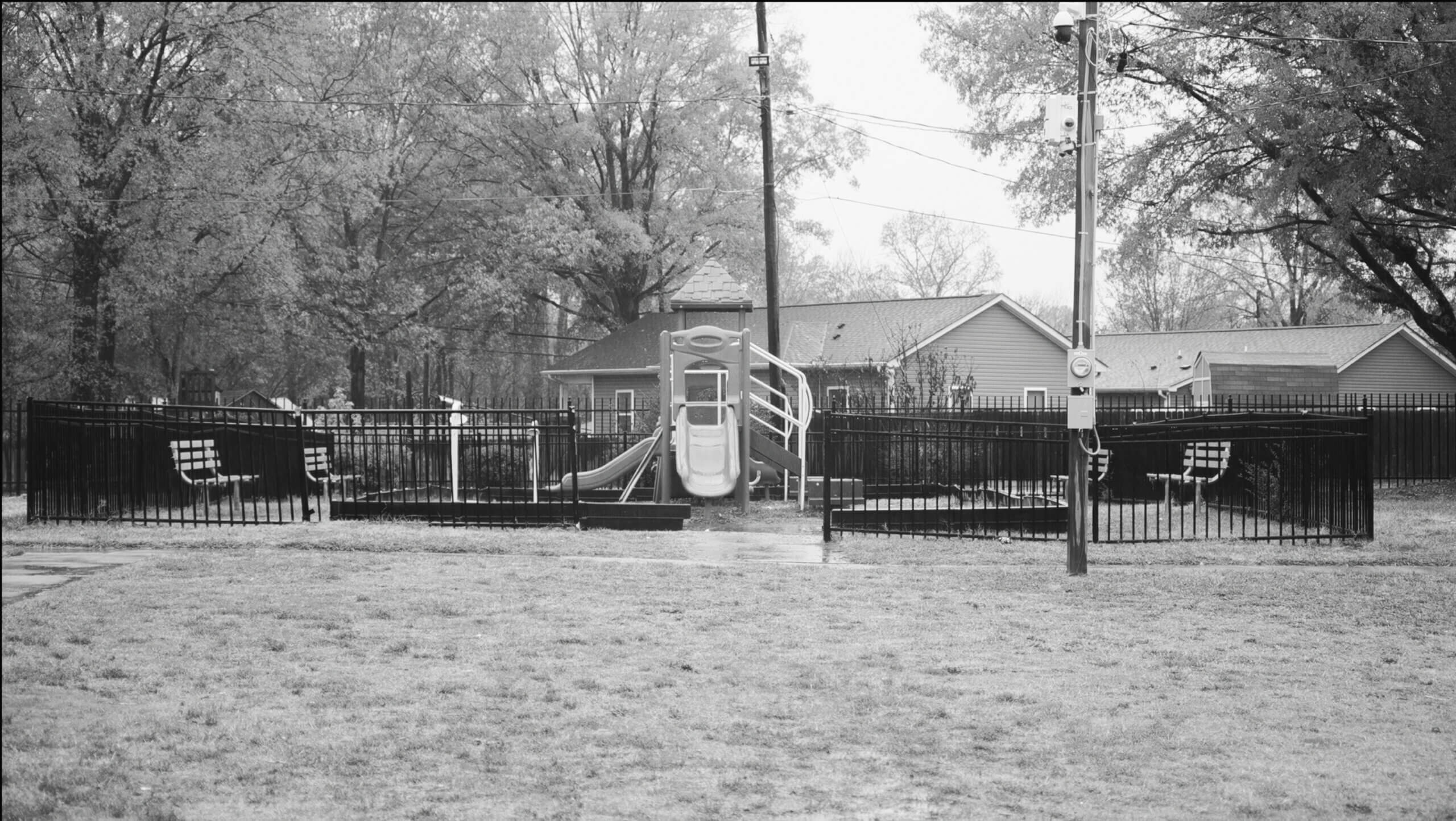 Black and white photo of an empty playground, on a rainy day. Surrounded by trees and grass