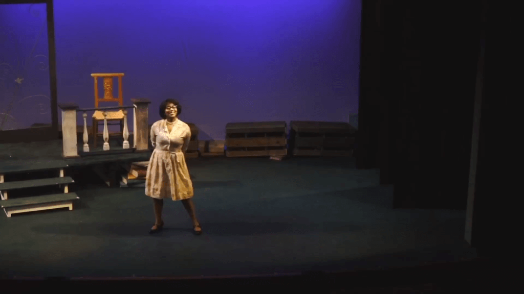 A younger adult black woman smiles on a stage. She is wearing a 1950s-style shirt and skirt and cat-eye glasses. The set on stage is sparse, with just a porch, and is dimly lit, as though for evening.