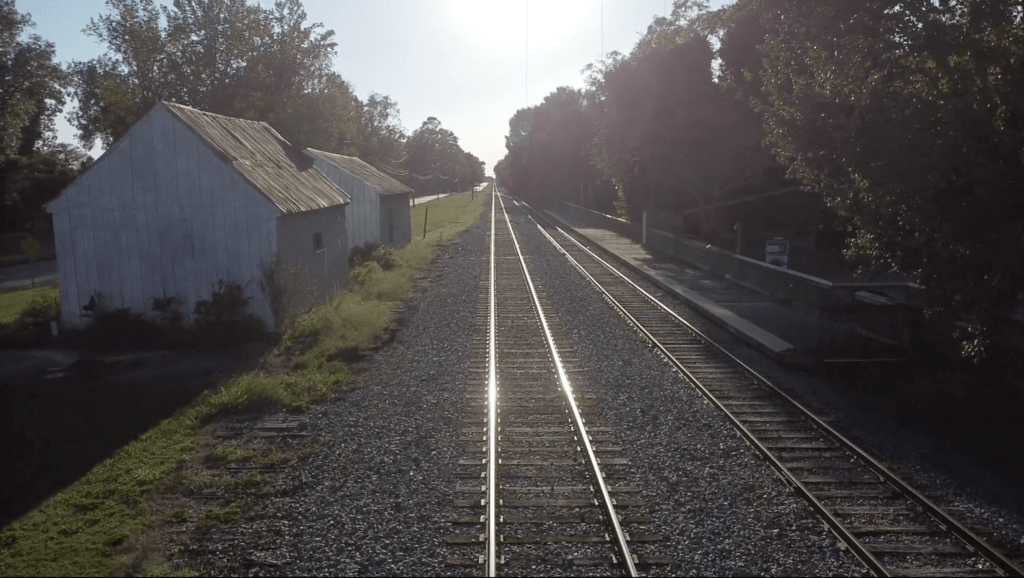 a set of train tracks with a rustic, rural setting featuring old buildings and a tree-lined path. The sunlight is shining directly on the tracks, creating a reflective glow.